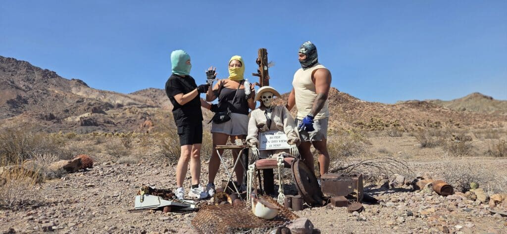 In Nevada’s desert near Las Vegas, masked figures gather by a skeleton at a quirky desk—perfect for Ghost Town sightseeing.