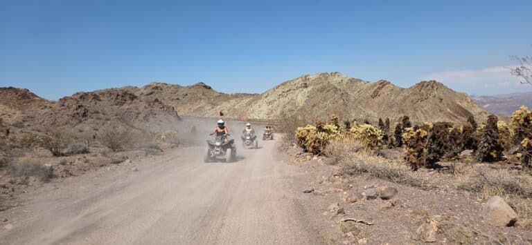 ATV riders explore a dusty desert trail near Las Vegas, Nevada, with rocky hills and cacti under a clear blue sky.