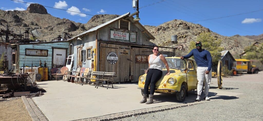 Two people pose by a yellow car at a Nevada ghost town near Las Vegas, with old signs and mountains, perfect for ATV tours.