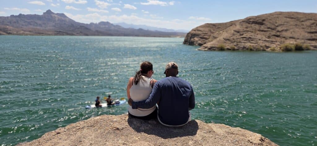 Couple on rocky ledge overlooks Colorado River near Nevada; paddlers below, mountains and blue sky create a peaceful Las Vegas scene.