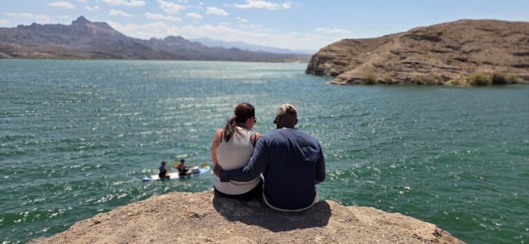 Couple on rocky ledge overlooks Colorado River near Nevada; paddlers below, mountains and blue sky create a peaceful Las Vegas scene.