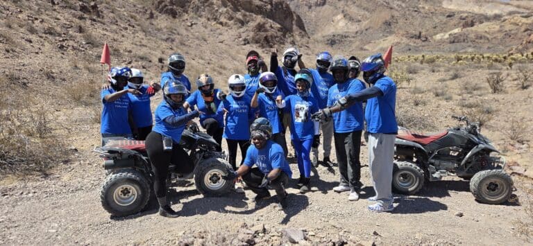 Smiling group in blue shirts on Las Vegas ATV tour, posing with helmets and ATVs in Nevada desert near Colorado River.