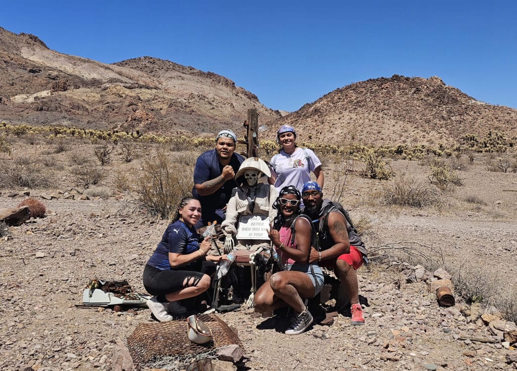 Group with skeleton in clothes by END ROUTE 66 sign, Nevada desert near Las Vegas mountains—perfect for Ghost Town sightseeing.