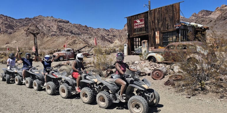 Helmeted riders on ATVs explore a Nevada ghost town near Las Vegas, passing vintage cars and rocky hills under blue skies.