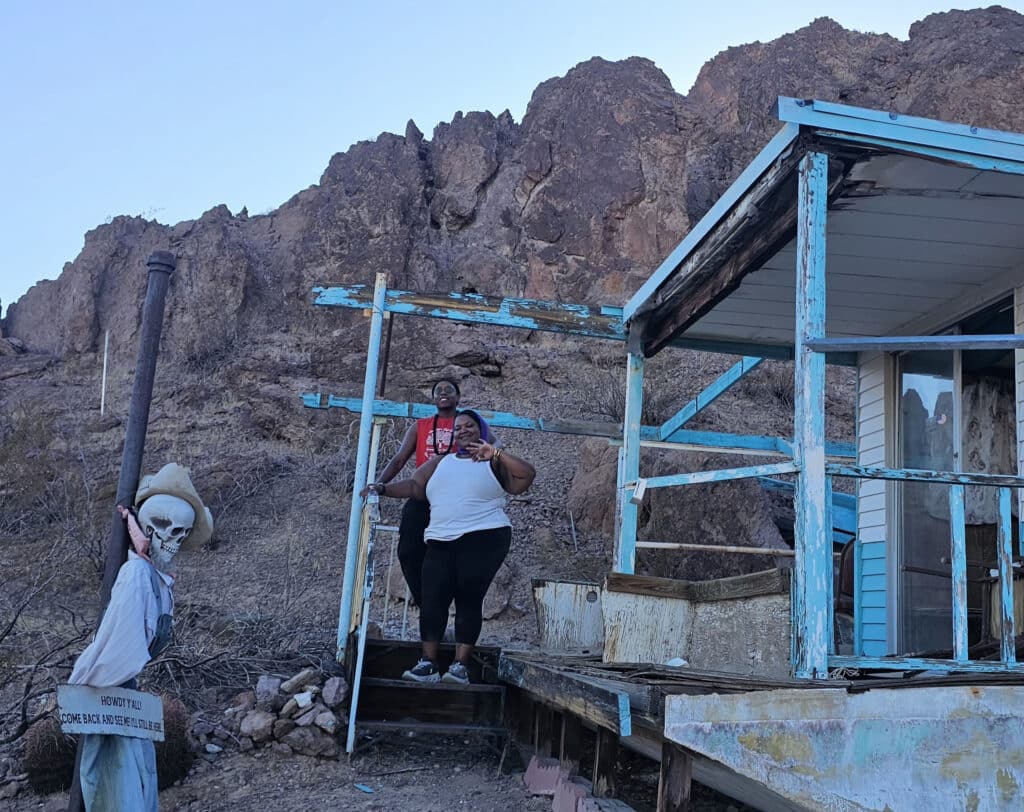 Two women smile on a ghost town deck near Las Vegas, Nevada; skeleton decor and rocky hills highlight desert sightseeing.