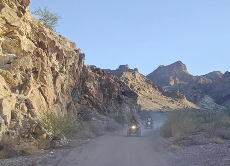 Adventurers on an ATV tour ride through a dusty Nevada desert canyon near Las Vegas with rugged cliffs and sparse vegetation.