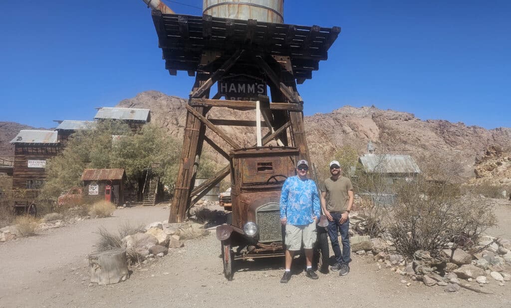 Smiling men by a vintage truck at Nevada ghost town near Las Vegas, with HAMMS tower—perfect for ATV and RZR desert tours.