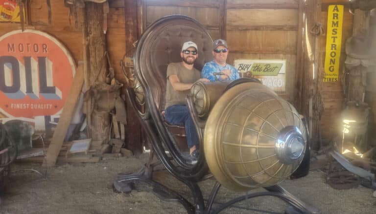 Two men smile on a steampunk chair in a vintage Las Vegas ghost town shed—perfect for Nevada ATV or RZR off-road adventures.
