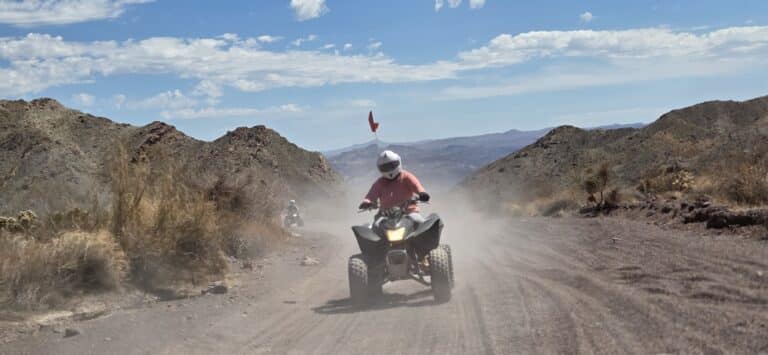 Rider in red shirt and helmet drives ATV on a dusty Nevada trail near Las Vegas, with mountains and another ATV behind.