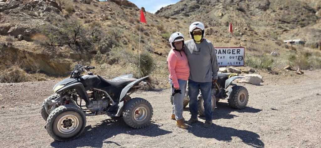 Riders in helmets stand by parked ATVs on a Nevada desert trail near Las Vegas, with danger sign and blue sky in the background.