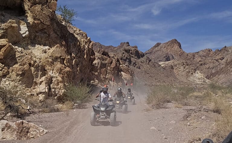 Adventurers ride ATVs on a dusty Nevada desert trail near Las Vegas with rugged mountains and sparse canyon views.