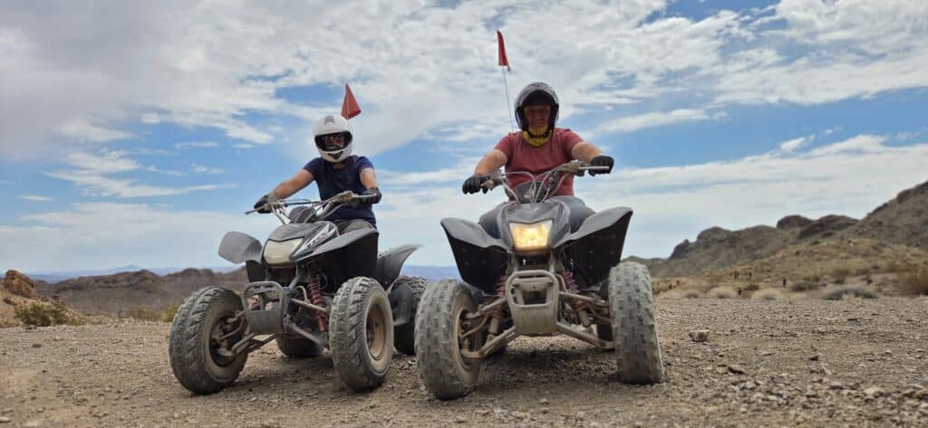 Riders with helmets enjoy an ATV tour near Las Vegas on a rocky desert trail. Both ATVs sport red flags under a partly cloudy sky.