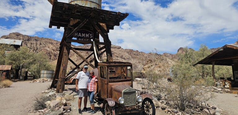 Couple by a rusty vintage car and “HAMM’S BEER” water tower in Nevada ghost town near Las Vegas; perfect for ATV tours.