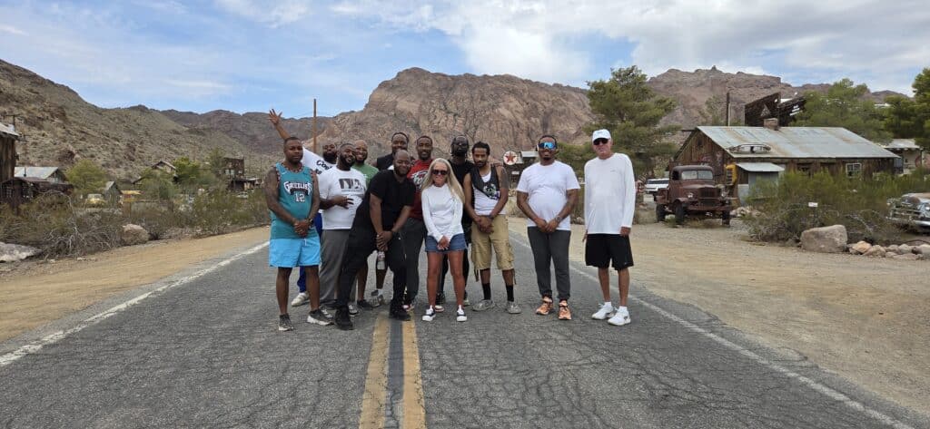 Twelve people pose on a Nevada ghost town road near vintage cars and rocky mountains, perfect for Las Vegas ATV or RZR adventures.