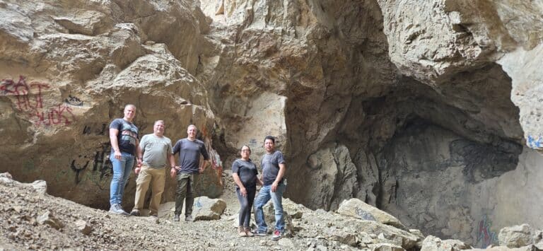 Group poses at Nevada cave entrance with graffiti, rocky ground—perfect for Las Vegas ATV tours near the Colorado River.