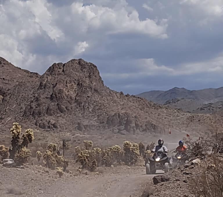 ATV riders explore a dusty trail near Las Vegas, Nevada, with cacti and rocky mountains under a partly cloudy sky.
