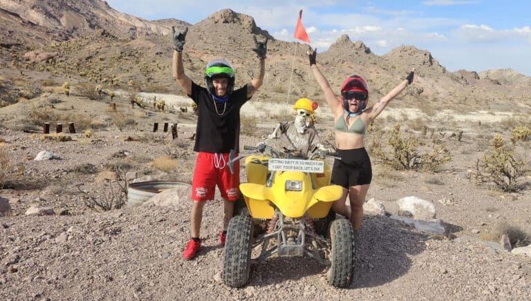 ATV tour near Las Vegas: two helmeted riders pose by a yellow ATV with skeleton driver in Nevada desert, mountains behind.