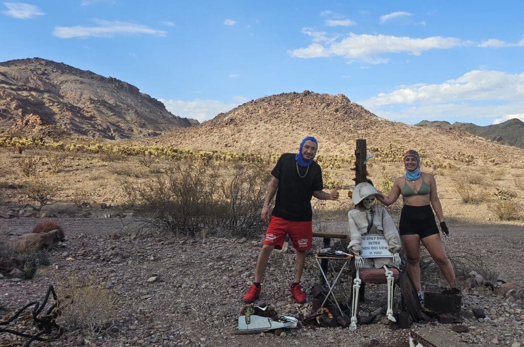 Two pose in a Nevada desert near Las Vegas; skeleton at ghost town desk, mountains and sparse brush under sunny sky, perfect for ATV tours.