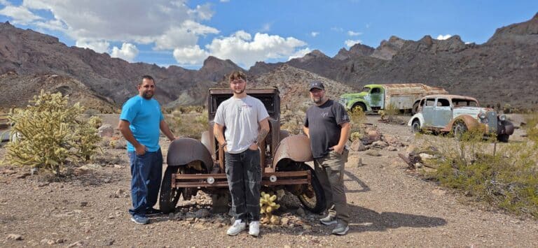 Three men pose by a rusty car in a Nevada desert near Las Vegas, surrounded by vintage vehicles and cacti—Ghost Town sightseeing.