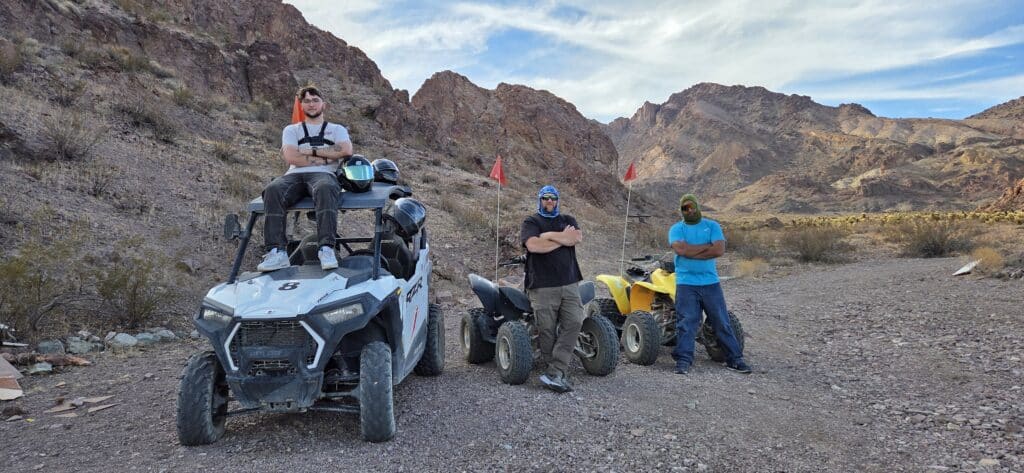 Three men with ATVs in Nevadas rocky desert—Las Vegas off-road adventure, mountains and blue sky in the backdrop.