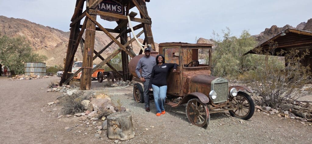 Smiling couple by a rusty vintage truck in Nevada desert near Las Vegas Ghost Town, mountains, and old wooden buildings.
