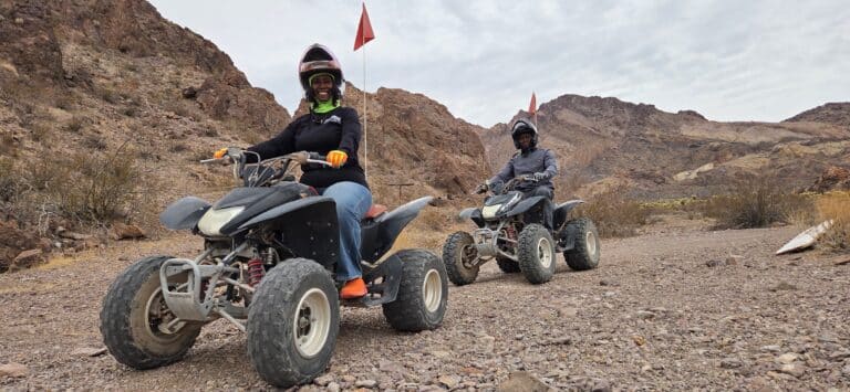 Helmeted riders on ATVs explore a rocky Nevada desert trail near Las Vegas, each ATV with a red flag and mountain views.