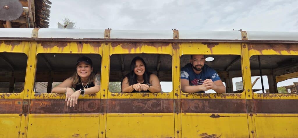 Three people smile from a vintage yellow bus during a Nevada Ghost Town sightseeing tour near the Colorado River.