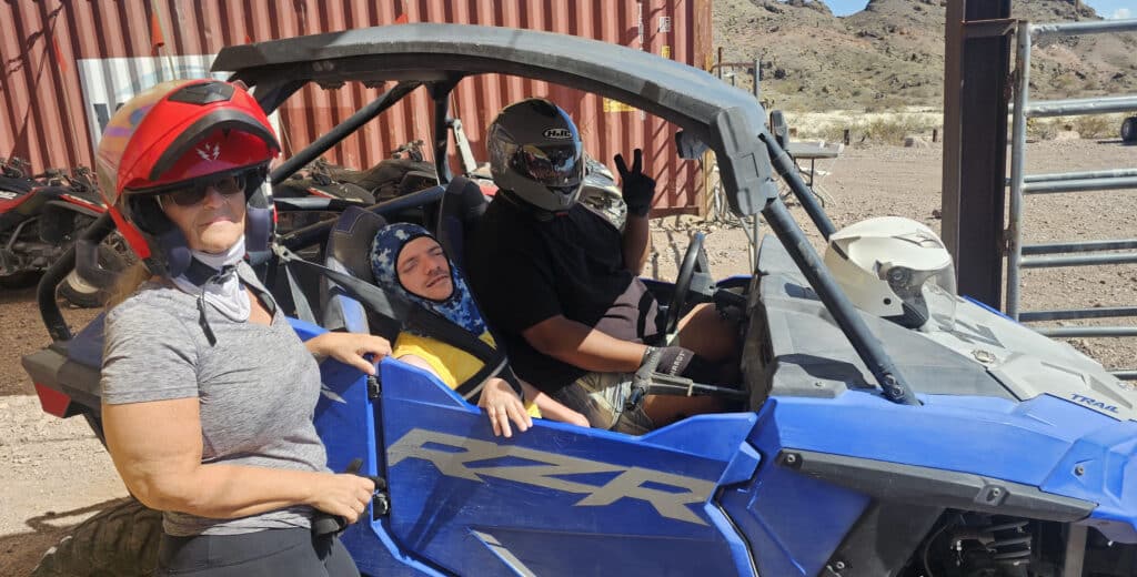Three on a Las Vegas ATV tour: woman by blue RZR, two men inside, all geared up, desert backdrop near Nevada’s Colorado River.