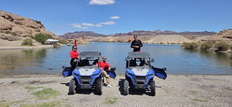 Adventurers in red shirts enjoy RZR off-road rides by the Colorado River near Las Vegas, Nevada, amid rocky hills and blue skies.