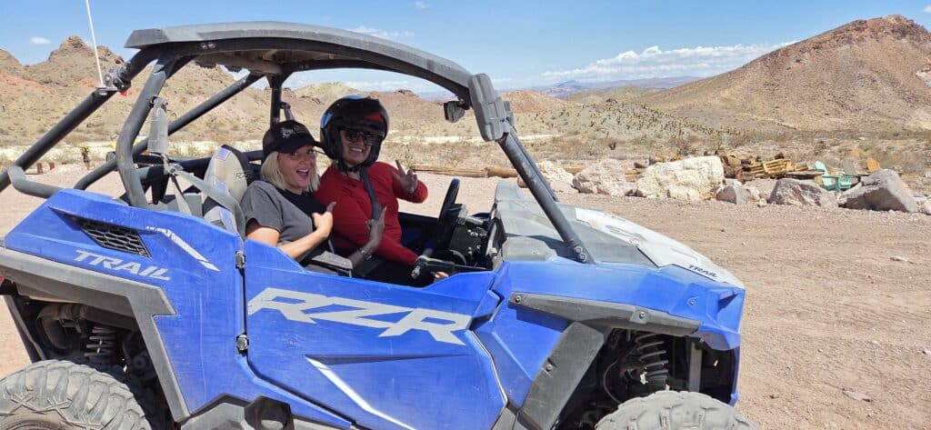 Two people smiling in a blue RZR on a Nevada desert ATV tour near Las Vegas, mountains and clear sky in the background.