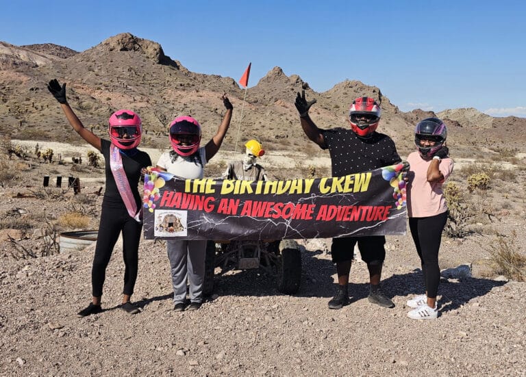 Four friends in helmets with a “Birthday Crew” banner on a Nevada ATV tour near Las Vegas, rocky hills and blue sky behind.
