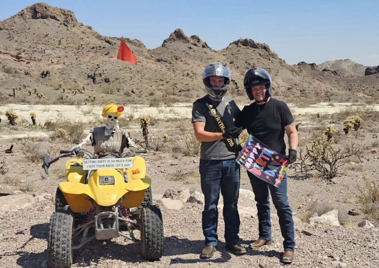 Two men in helmets shake hands in a Nevada desert; a skeleton on an ATV. Perfect for Las Vegas ATV tours and Ghost Town sightseeing.
