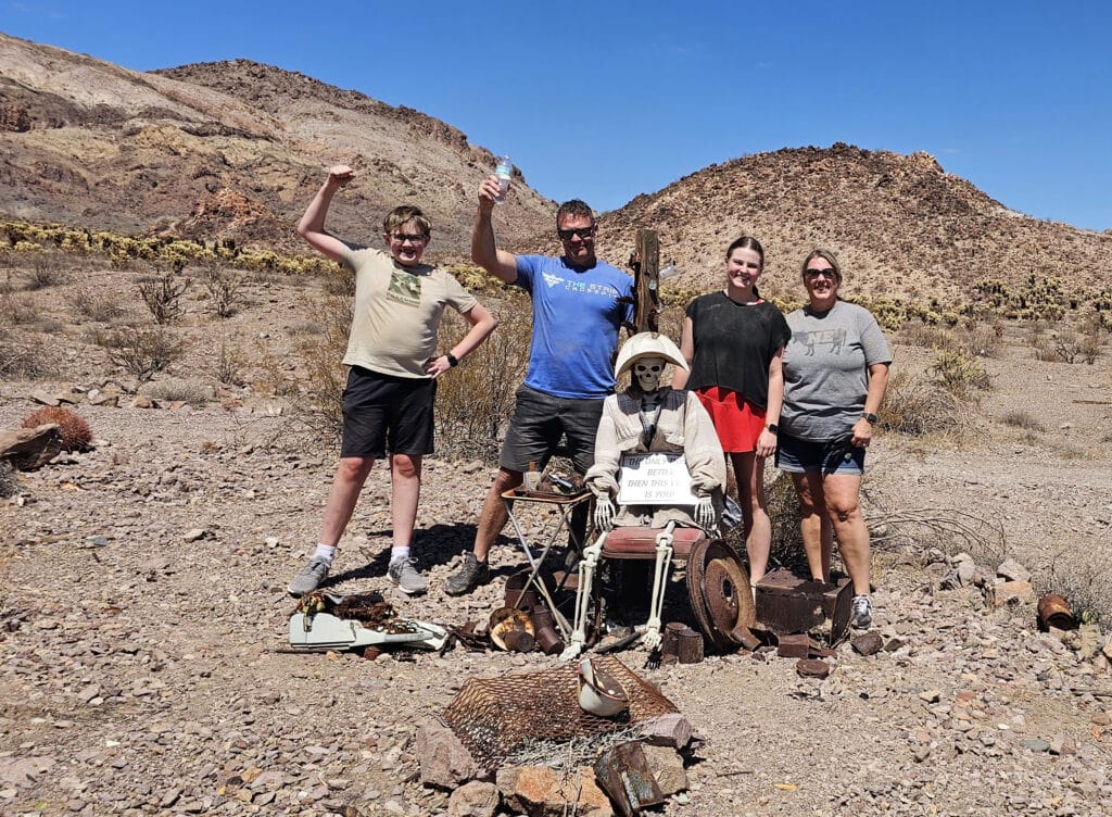 Friends smile by a dressed skeleton on old machinery in a Nevada desert ghost town near Las Vegas under sunny skies.