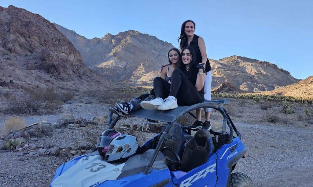 Three women smile atop a blue ATV in Nevada desert near Las Vegas, ready for RZR off-road tours by the Colorado River.