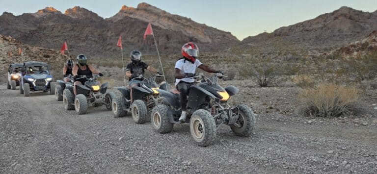 ATV tour near Las Vegas: four riders with red flags on a desert trail, RZR following, Nevada mountains under clear sky.