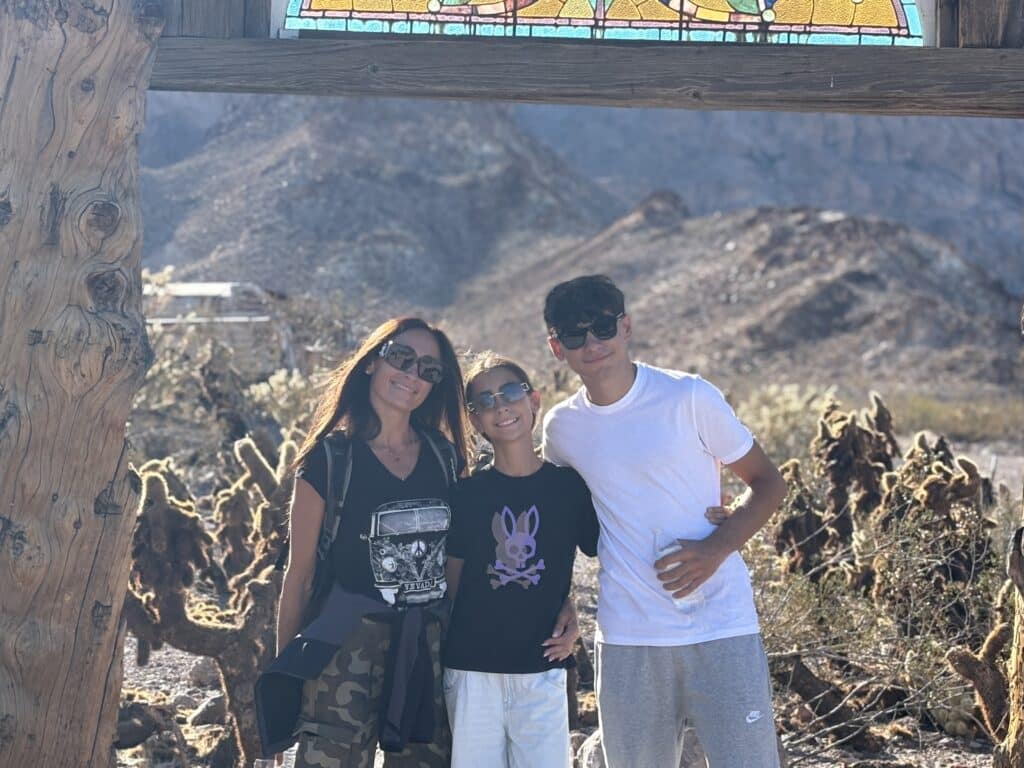 A woman and two teens in sunglasses smile under a wooden arch near Las Vegas with cacti, mountains, and desert ATV tours.