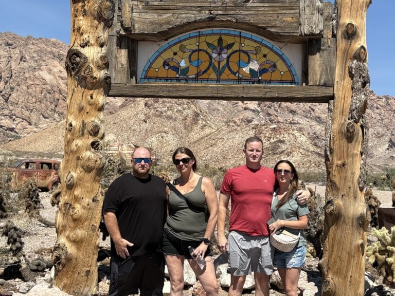 Four adults pose under a rustic wooden arch in a Nevada desert near Las Vegas, with mountains, an old car, and Ghost Town views.