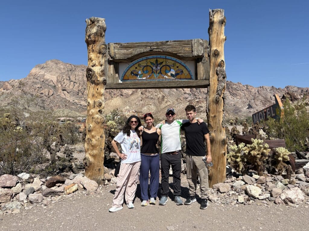 Group stands arm-in-arm under a wooden sign in the Nevada desert near Las Vegas, with cacti, mountains, and clear blue sky.