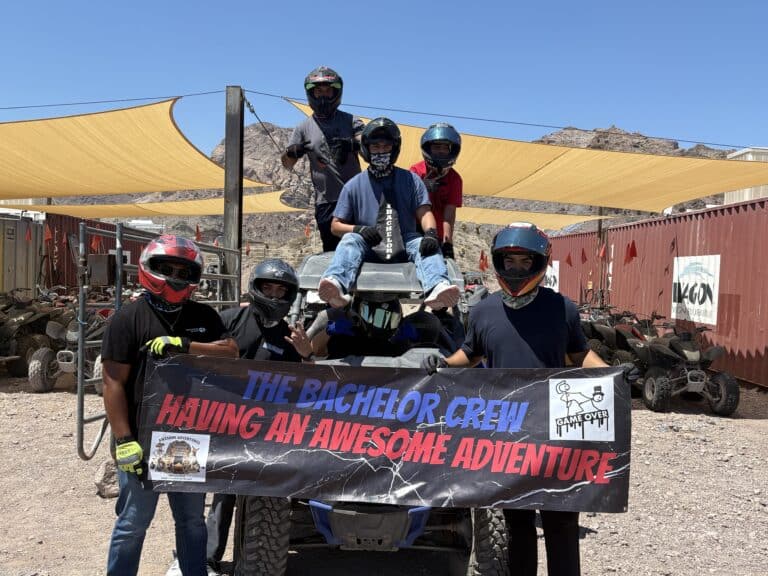 Group in helmets with ATV holds “The Bachelor Crew Having an Awesome Adventure” banner at Nevada ATV tour site near Las Vegas.