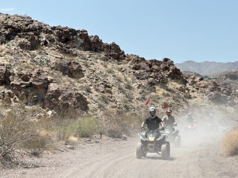 Adventurers ride ATVs on a dusty desert trail near Las Vegas, Nevada, with rocky hills—perfect for ATV tours or Ghost Town sightseeing.