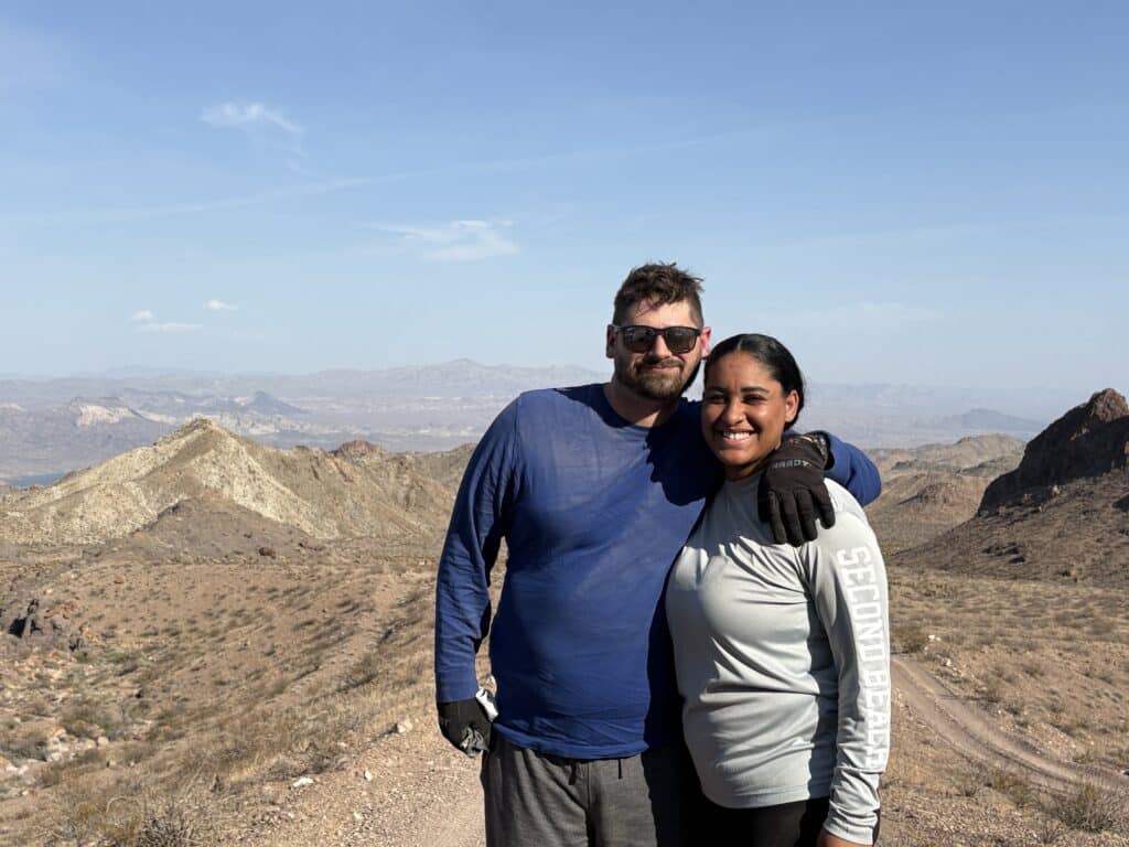 Two people in gloves and long sleeves smile on a Nevada desert trail near Las Vegas with mountains, ideal for ATV tours.
