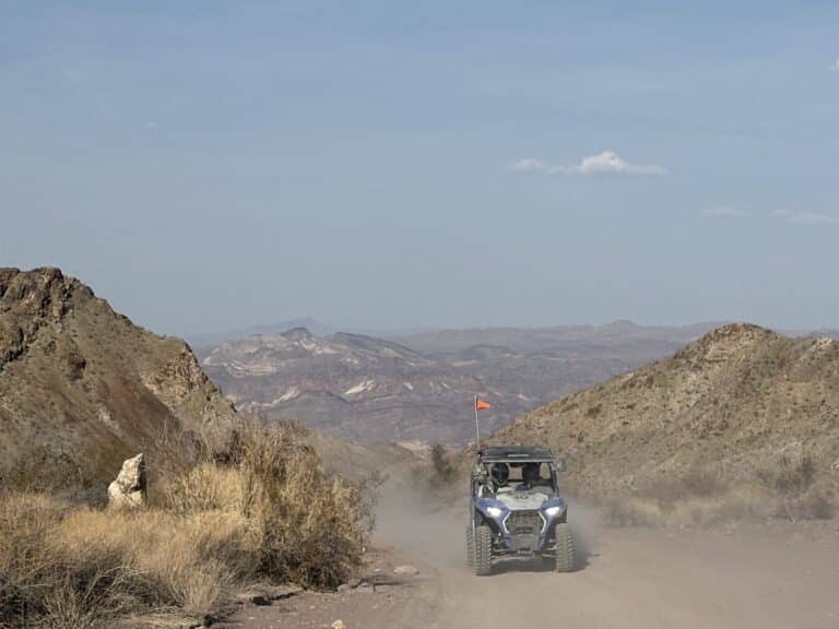 Blue RZR off-road vehicle kicks up dust on a Nevada desert trail near Las Vegas, with rocky hills and mountains in the background.