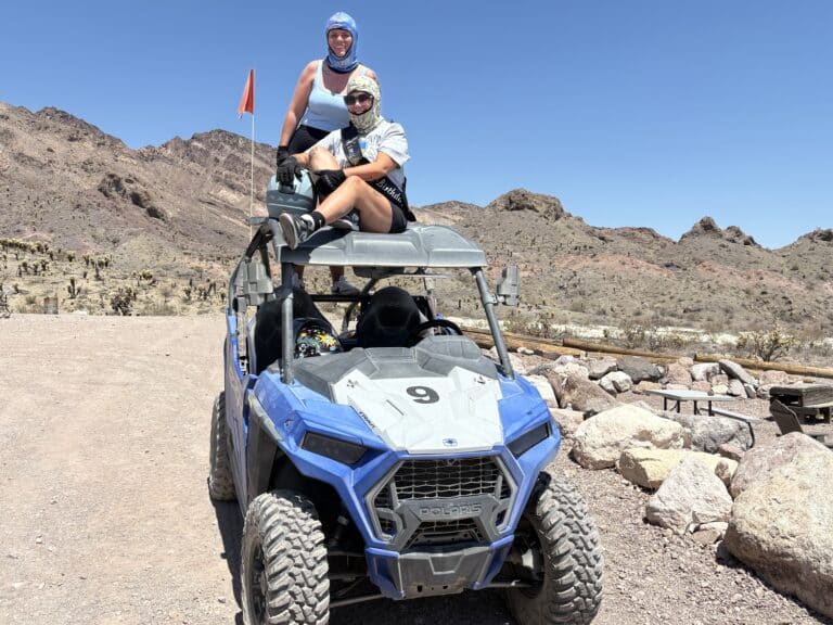 Two people smile atop a blue Polaris RZR on a Nevada desert ATV tour near Las Vegas under a clear sky.
