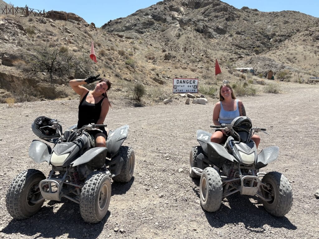 Smiling women on ATVs pose in Nevada’s rocky desert near Las Vegas with “Danger” sign—perfect for ATV tours or off-road adventures.