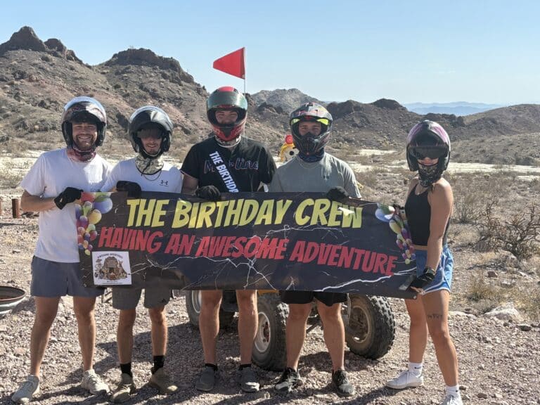 Group in helmets pose with The Birthday Crew sign during a Las Vegas ATV tour, quad and Nevada desert hills behind them.