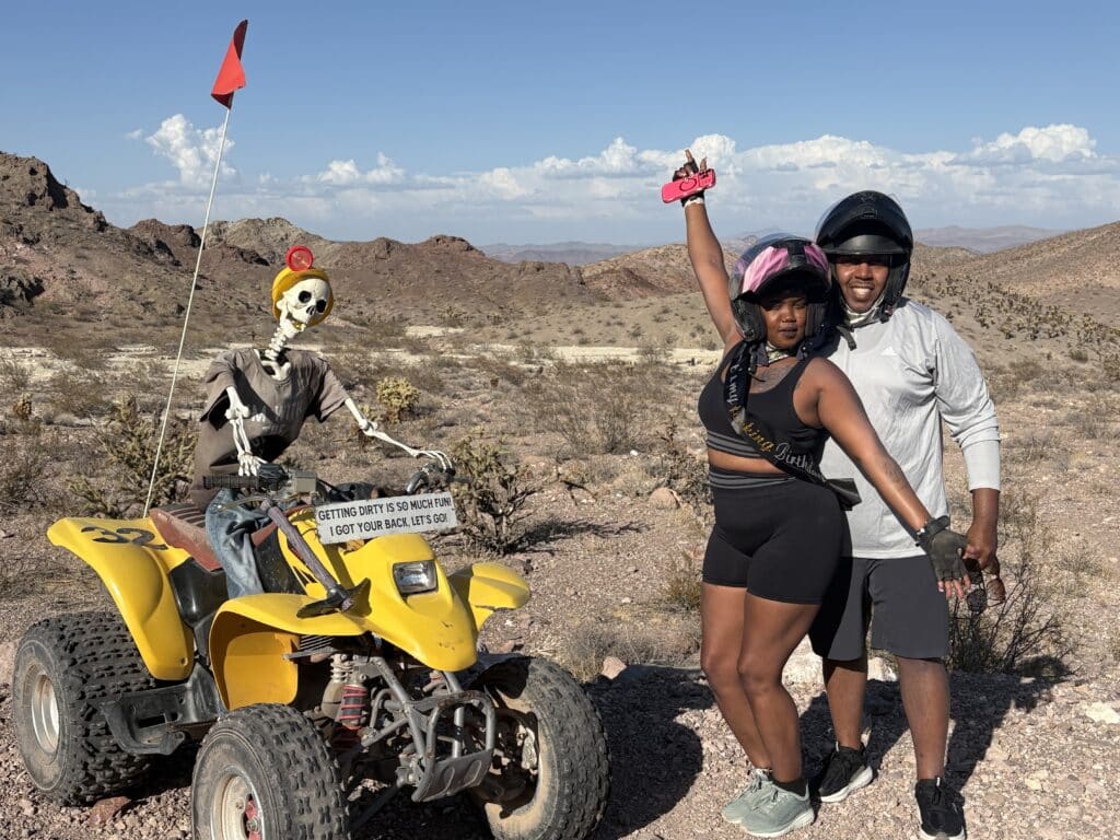 Smiling couple in helmets poses near a yellow ATV with skeleton rider, Las Vegas desert ATV tour, rocky Nevada hills behind.