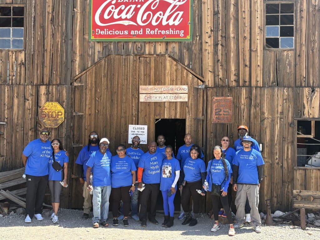 Smiling group in blue shirts at a rustic Nevada ghost town building, vintage Coca-Cola sign, sunny day—perfect for ATV tours near Las Vegas.