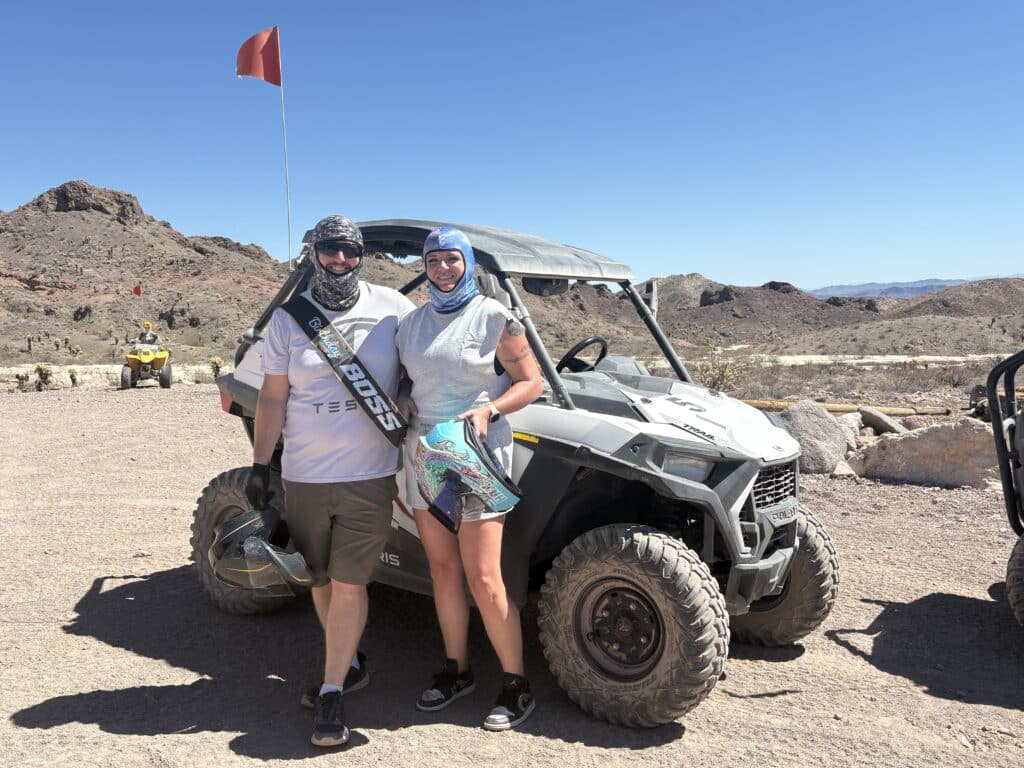 Adventurers in protective gear pose by an ATV with a red flag in Nevada desert near Las Vegas, rocky terrain and mountains behind.