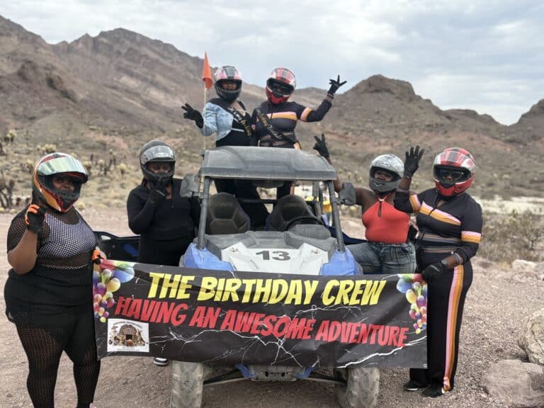 Six friends in helmets and bright gear pose by an ATV near Las Vegas, enjoying a birthday RZR tour in the Nevada desert.