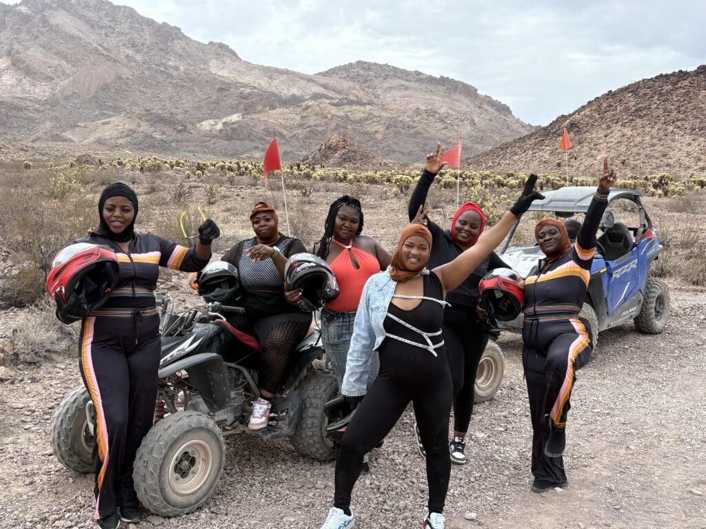 Six women celebrate in sporty gear by ATVs amid Nevada desert hills, enjoying a thrilling Las Vegas ATV tour adventure.